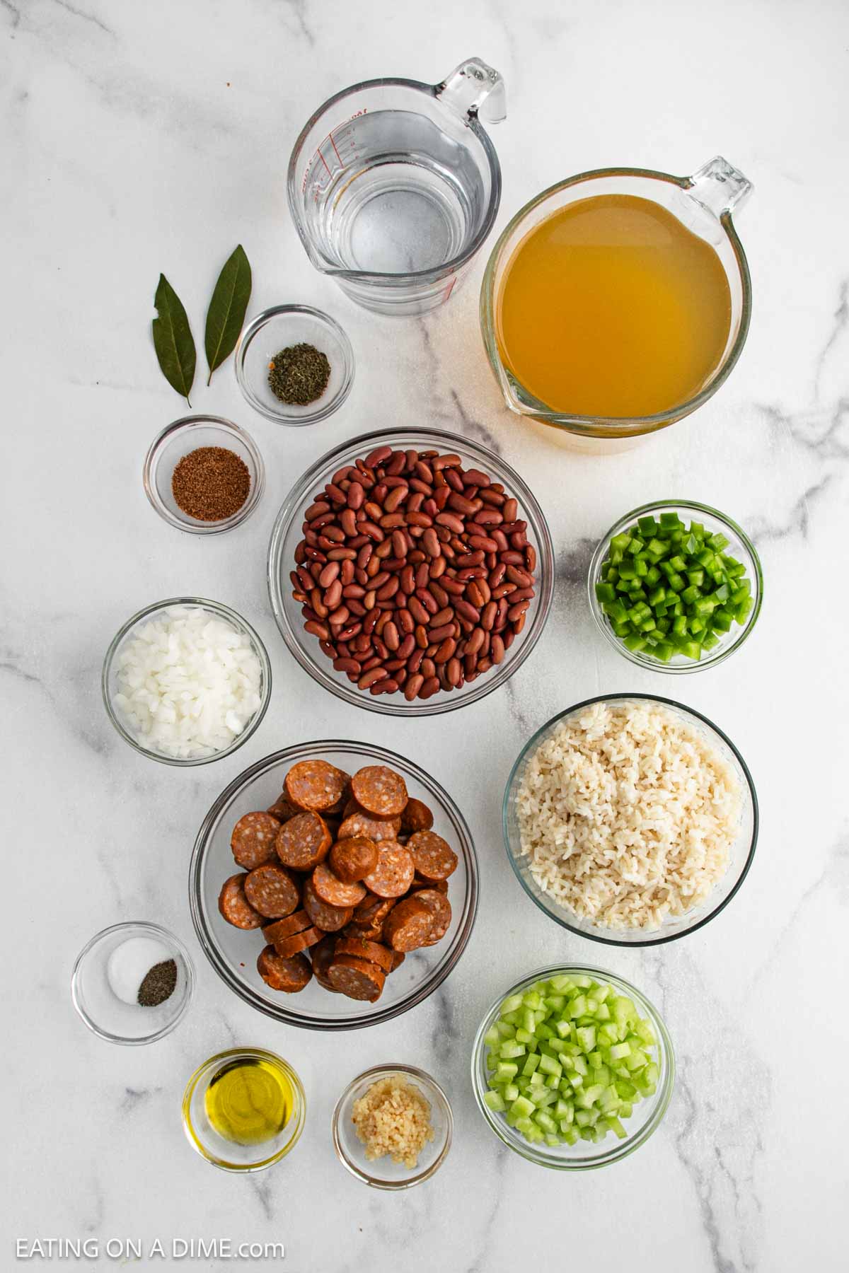 Overhead view of ingredients for Red Beans and Rice, including sliced sausage, cooked rice, kidney beans, chopped onions, green pepper, celery, garlic, bay leaves, spices, olive oil, water, and chicken broth on a white surface.