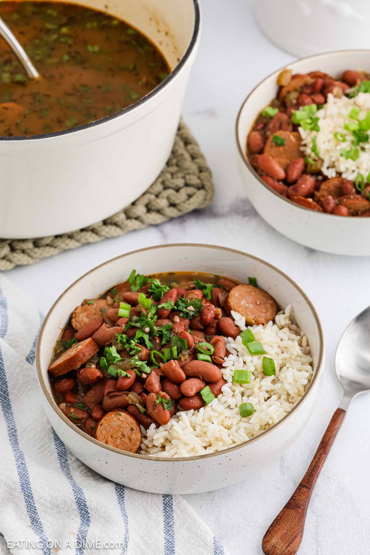 A bowl of Red Beans and Rice features sliced sausage and chopped green onions, served with white rice. A spoon rests nearby, with a pot of more red beans in the background.
