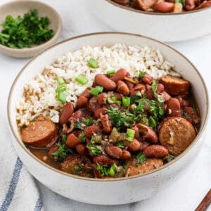 A bowl of classic Red Beans and Rice features white rice topped with red beans and sausage slices, garnished with chopped green onions and parsley. A small dish of fresh herbs sits nearby.