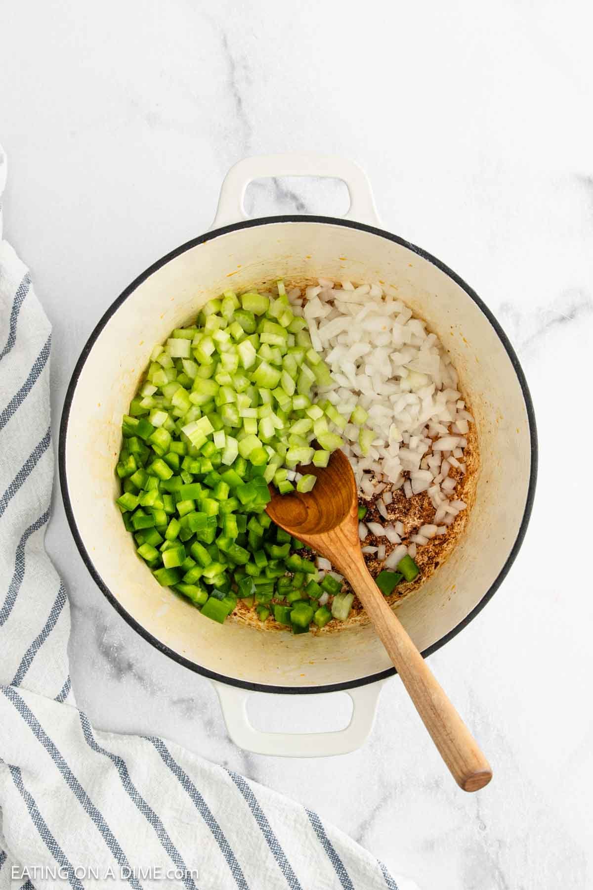 A white pot on a marble surface contains chopped celery, green bell pepper, and onion—classic ingredients for Red Beans and Rice. A wooden spoon rests inside the pot, with a striped towel nearby.