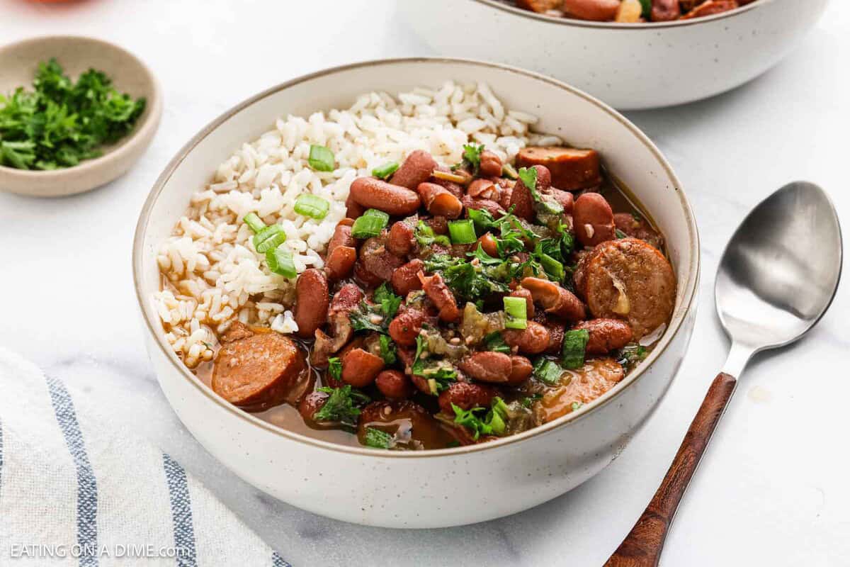 A bowl of Red Beans and Rice topped with sliced sausage and green onions, garnished with fresh herbs. A spoon rests beside the bowl, and another serving of this classic Red Beans dish is visible in the background.
