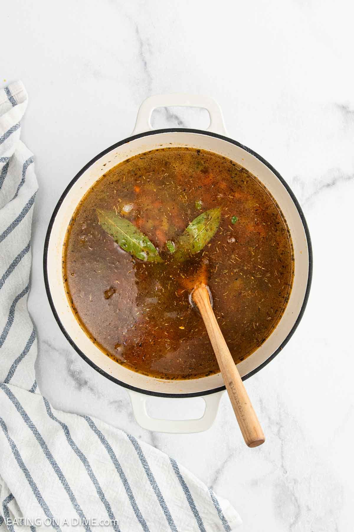 A white pot filled with dark broth, herbs, and two bay leaves, ideal for making Red Beans and Rice, sits on a marble surface with a wooden spoon and a striped dish towel nearby.