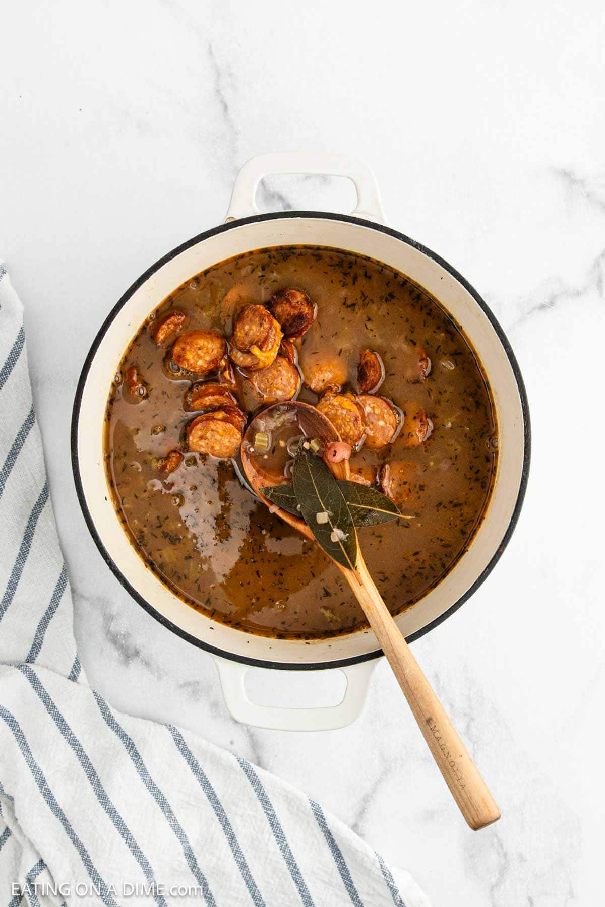 A pot of sausage gumbo in a white Dutch oven, with sliced sausage pieces in a dark, seasoned broth reminiscent of Red Beans and Rice. A wooden spoon with a bay leaf rests inside the pot, and a striped kitchen towel is nearby.