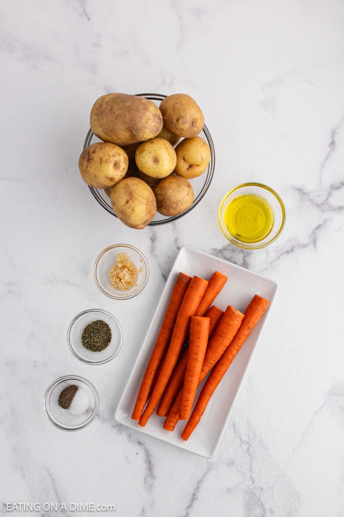 A bowl of whole potatoes, a plate with carrots, and small bowls of olive oil, minced garlic, salt, and pepper rest on a white marble surface—perfect for preparing roasted potatoes and carrots.