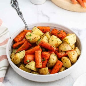 A white bowl filled with roasted potatoes and carrots, seasoned with herbs and garnished with chopped parsley, sits on a white surface with a silver spoon resting inside—an inviting medley of roasted vegetables.
