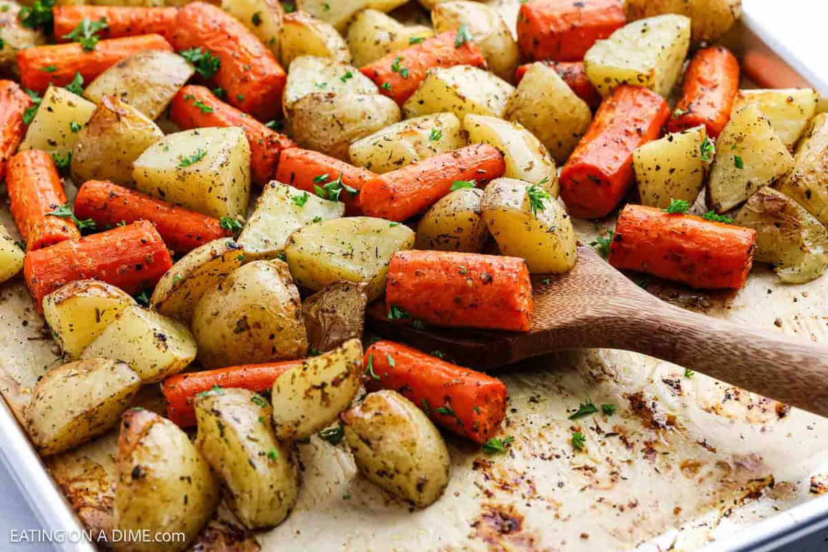 A wooden spoon scoops roasted carrots and potatoes, perfectly golden and seasoned with herbs, from a baking sheet. The roasted vegetables are garnished with freshly chopped parsley.