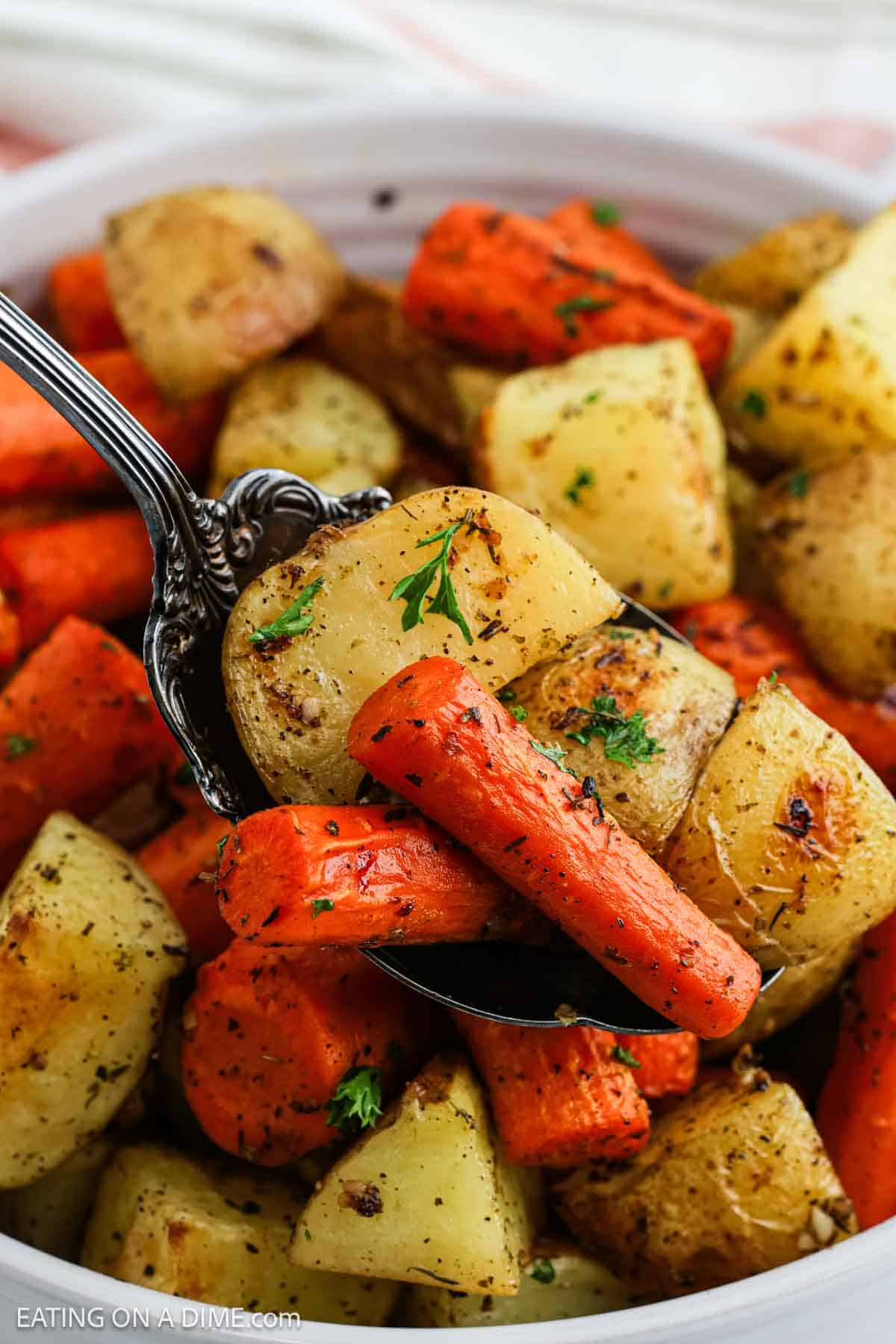 A close-up of roasted potatoes and carrots, seasoned with herbs, served in a bowl with a spoon lifting a portion. The roasted potatoes and carrots are golden brown and garnished with fresh parsley.