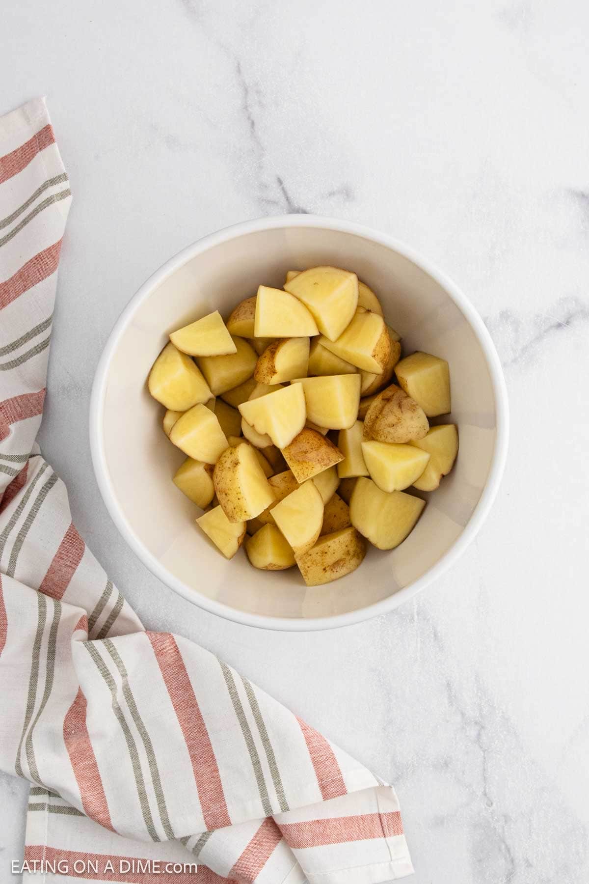 A white bowl filled with chopped potatoes sits on a white marble surface next to a striped cloth with beige, red, and gray lines, ready to be turned into delicious roasted potatoes.