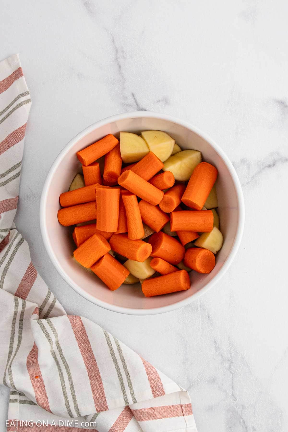 A white bowl filled with chopped roasted potatoes and carrots sits on a light marble surface next to a striped kitchen towel.
