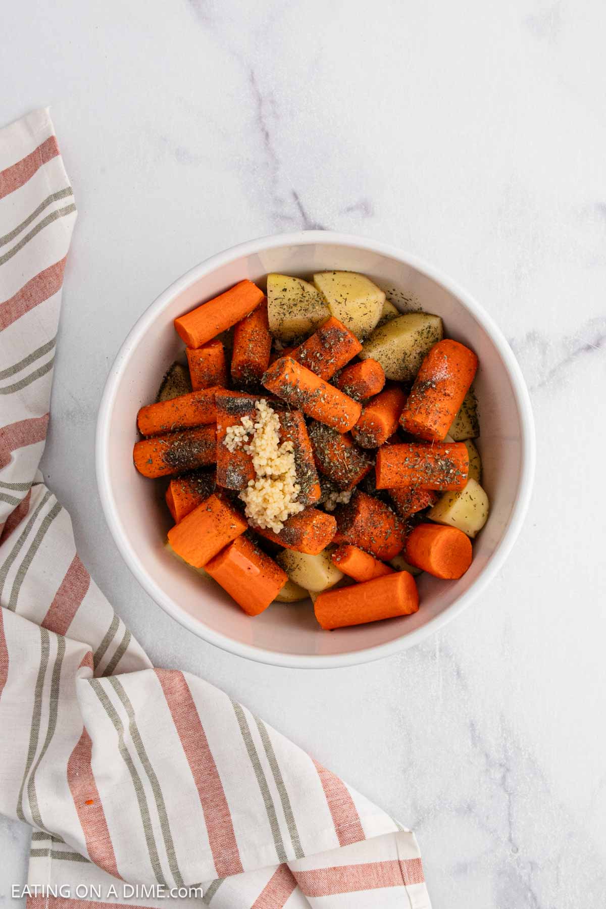A white bowl filled with roasted potatoes and carrots, topped with herbs and minced garlic, sits on a white marble surface next to a striped cloth napkin.
