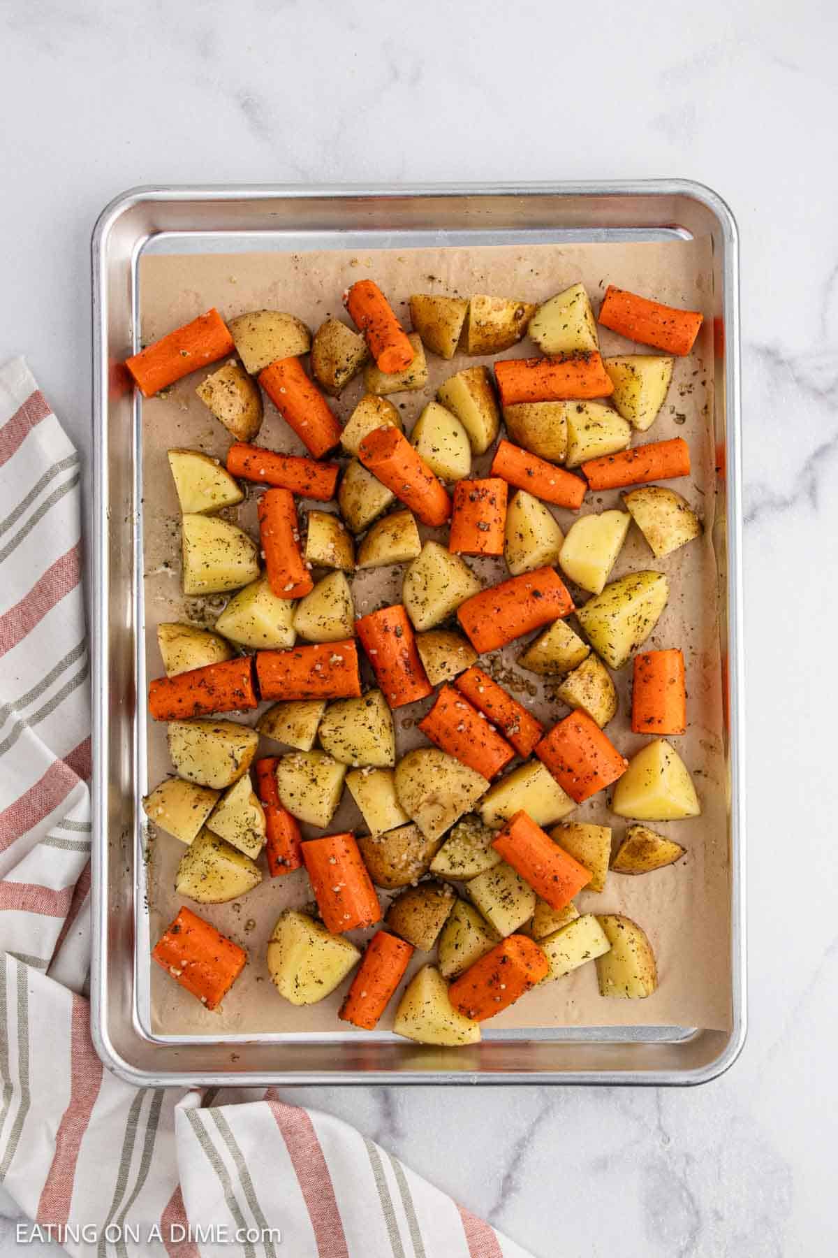 A baking sheet with roasted potatoes and carrots, seasoned with herbs, is ready for the oven. Lined with parchment paper, it sits on a marble surface beside a striped cloth, promising deliciously caramelized veggies.