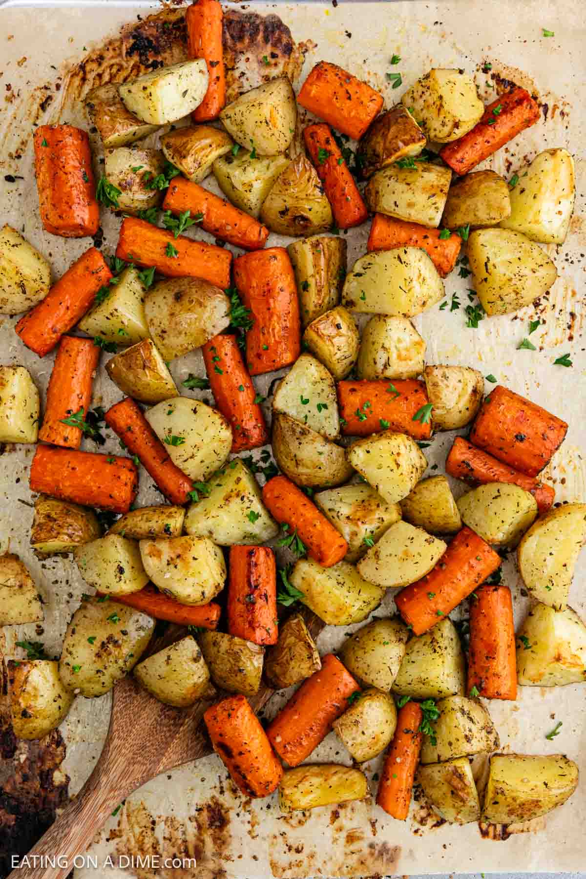 Roasted chunks of carrots and potatoes, classic roasted vegetables, seasoned with herbs, spread on a baking sheet with a wooden spoon and sprinkled with chopped parsley.