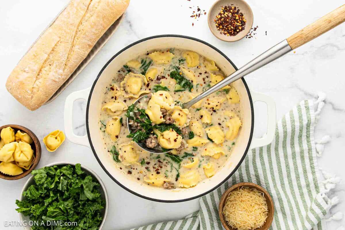 A white pot of creamy tortellini soup with spinach and red pepper flakes sits on a striped towel. A ladle is lifting the hearty sausage and tortellini soup, surrounded by bread, shredded cheese, and leafy greens.