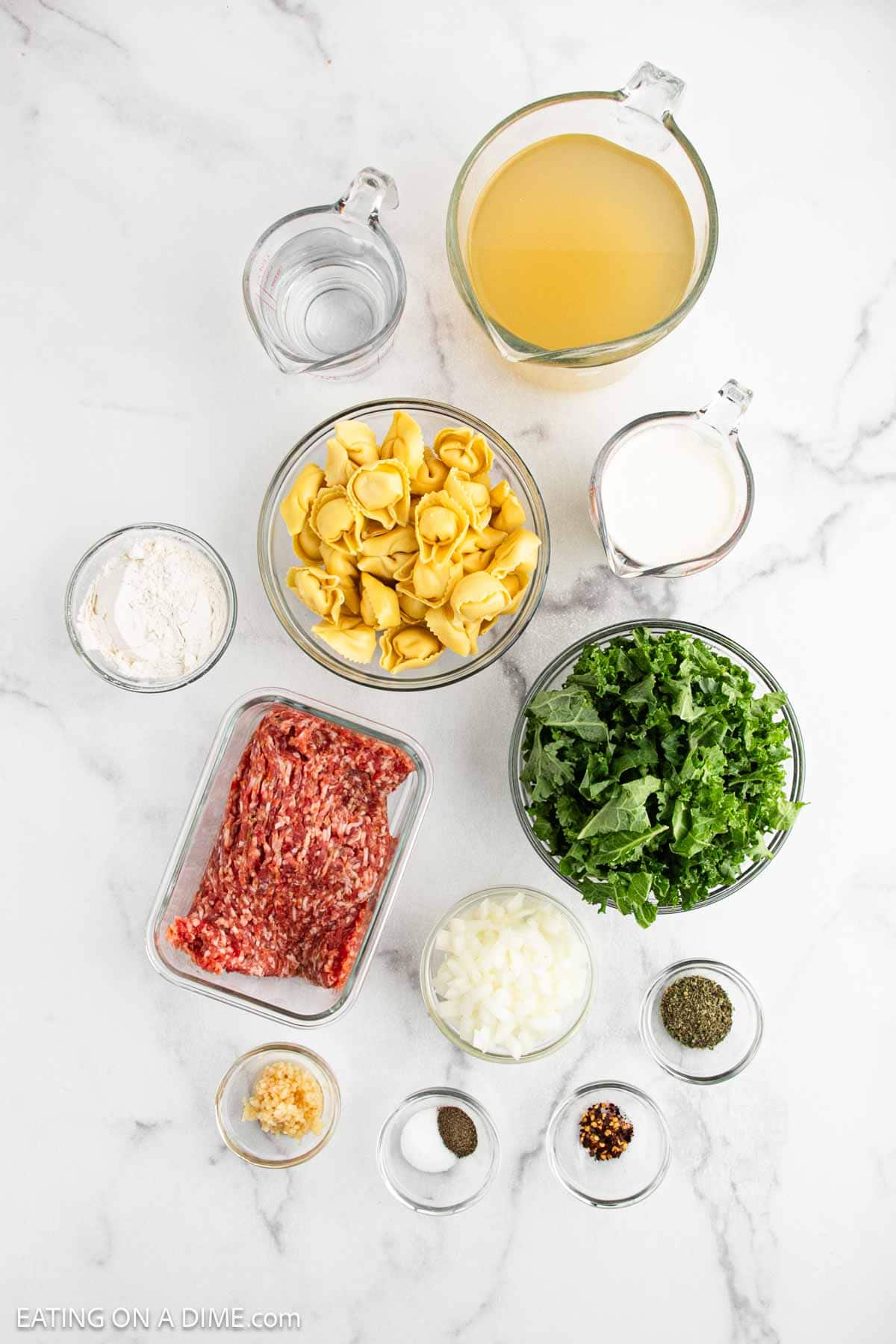 Top-down view of ingredients for Sausage Tortellini Soup on a white surface: ground sausage, chopped onion, minced garlic, tortellini pasta, chopped kale, flour, broth, milk, water, and small bowls of salt, pepper, herbs, and red pepper flakes.