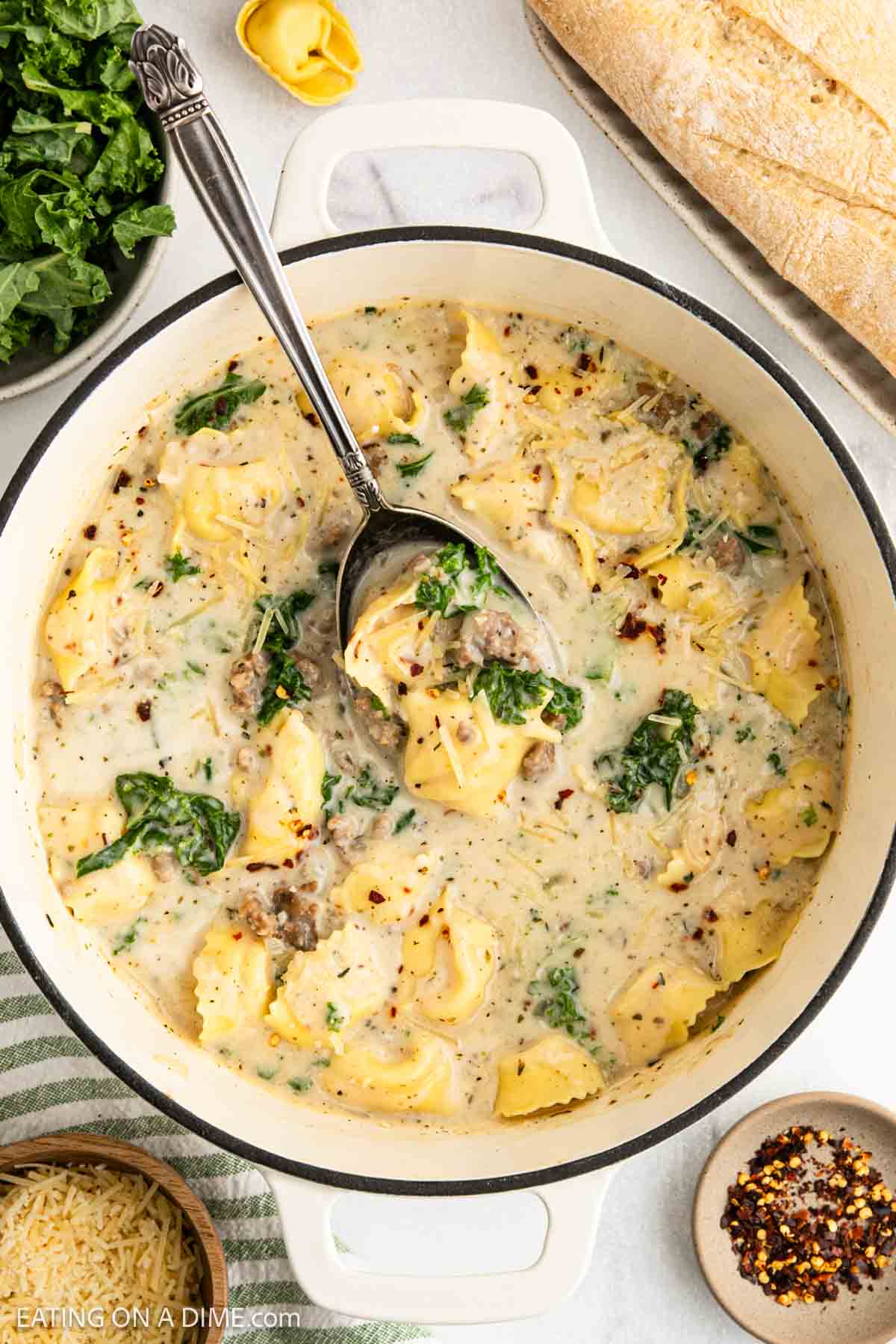 A pot of creamy tortellini soup with savory sausage and spinach, garnished with herbs. A spoon rests inside the pot. Bread, grated cheese, and a bowl of red pepper flakes are nearby on the table.
