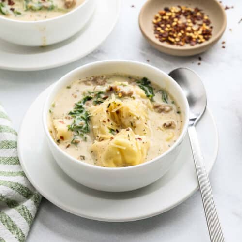 A bowl of creamy Sausage Tortellini Soup with sausage, spinach, and tortellini is served with a spoon on a white saucer. Red pepper flakes and another bowl of Tortellini Soup are in the background.