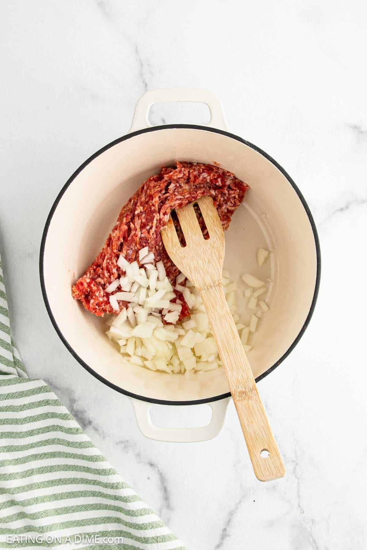 A white pot containing ground beef and chopped onions, with a wooden slotted spoon on top. Perfect as the savory base for a comforting sausage tortellini soup, it sits on a white marble surface next to a green and white striped towel.