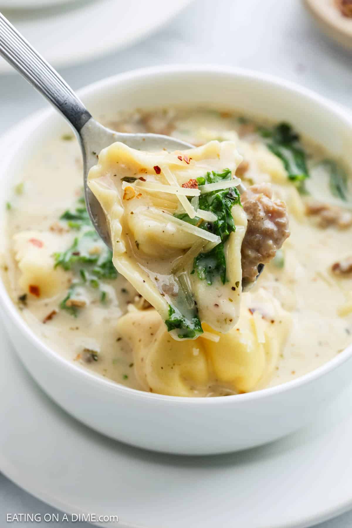 A close-up of a spoonful of creamy tortellini soup with sausage, kale, and shredded cheese being lifted from a white bowl brimming with comforting tortellini soup.
