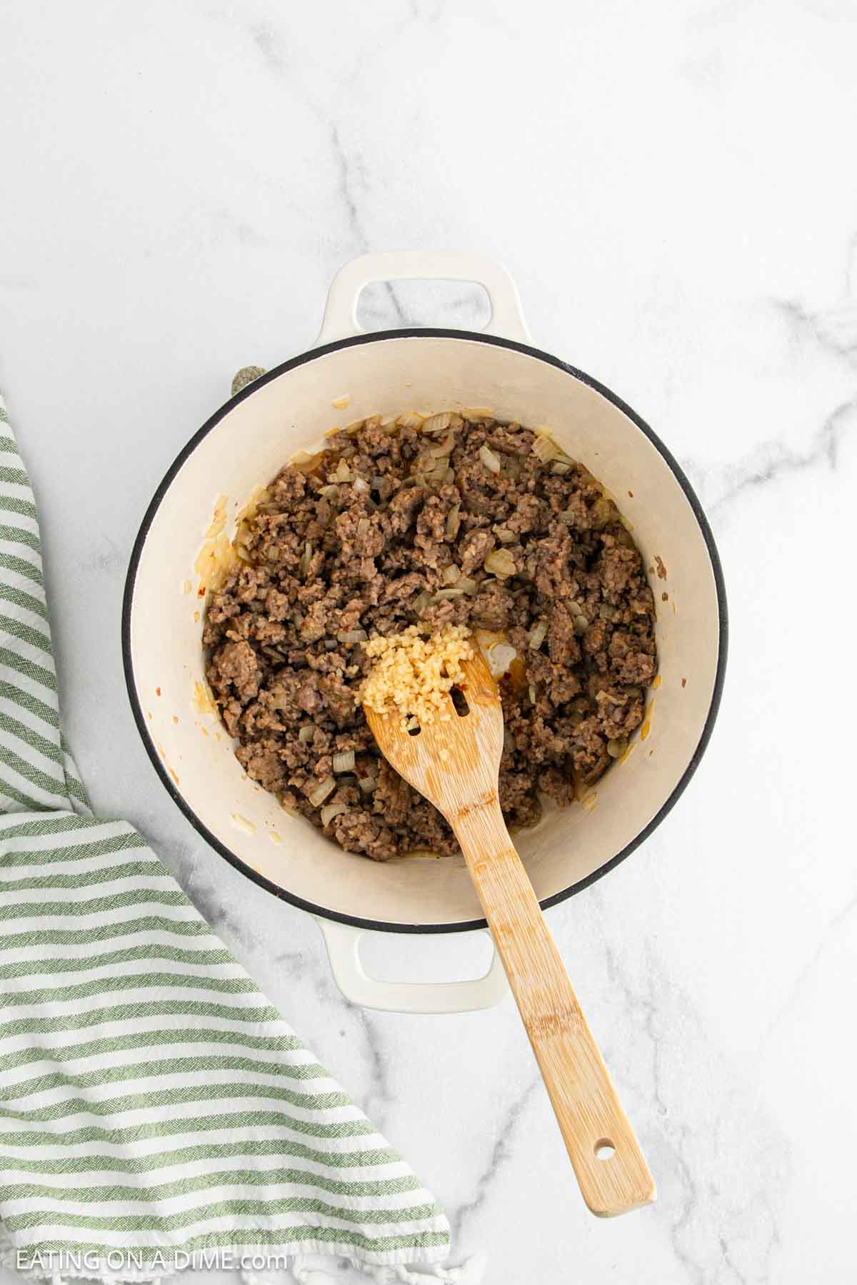 A white pot filled with cooked sausage, onions, and garlic being stirred with a wooden spoon. Perfect as a base for soup or tortellini dishes. A green and white striped kitchen towel rests to the side on a white marble surface.