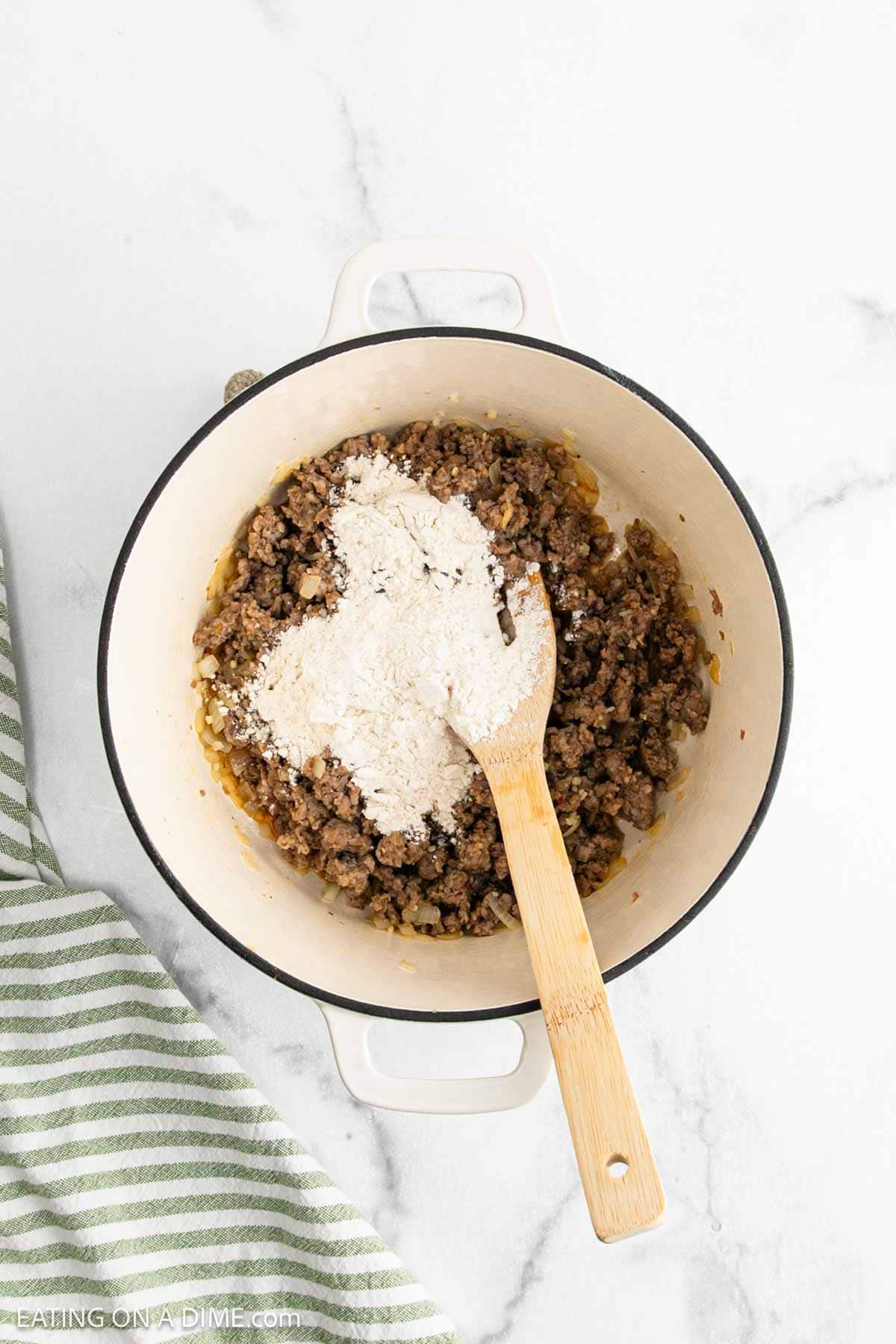 A white pot holds cooked sausage with a pile of flour on top, being stirred with a wooden spoon. A green and white striped towel is next to the pot on a white marble surface, perfect for starting Sausage Tortellini Soup.