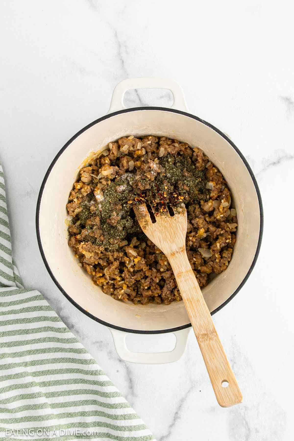A white pot filled with cooked sausage and spices being mixed with a wooden spoon, sitting on a marble countertop next to a green and white striped towel.