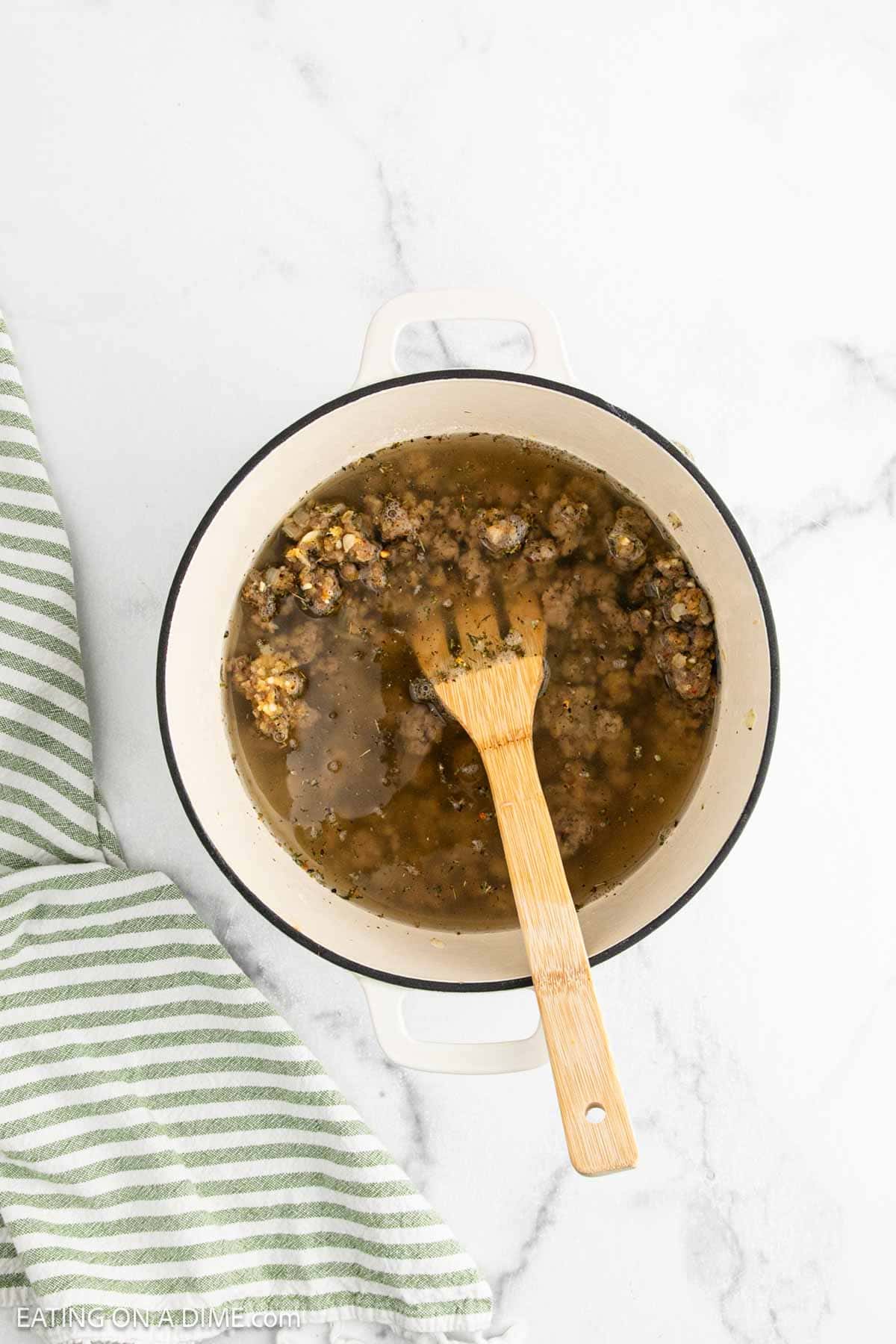 A white pot filled with Sausage Tortellini Soup sits on a white marble surface. A wooden spoon rests inside the pot, and a green-striped kitchen towel is placed nearby.