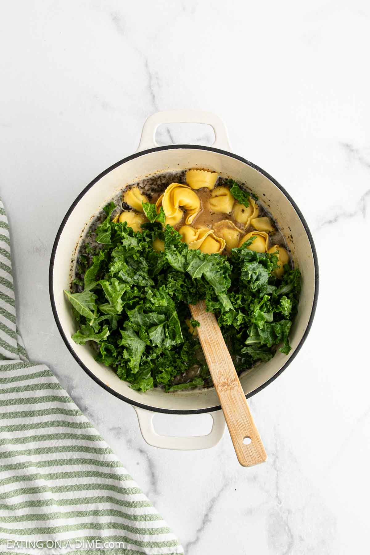 A white pot on a marble surface contains Sausage Tortellini Soup with yellow tortellini and fresh green kale, being stirred with a wooden spoon. A green and white striped towel lies beside the pot.
