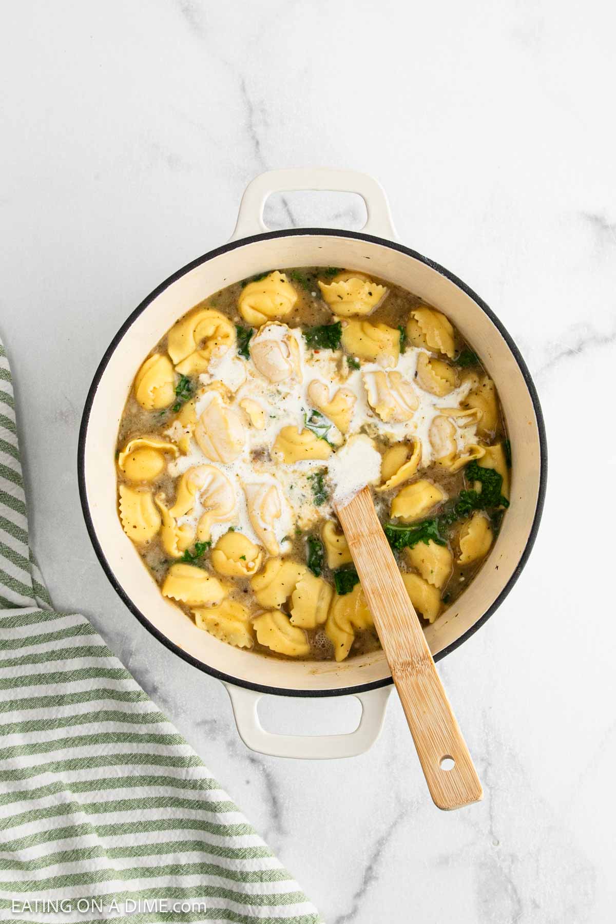 A white Dutch oven filled with tortellini soup, savory sausage, fresh spinach, and a swirl of cream sits on a marble countertop. A wooden spoon rests in the pot, with a green striped towel nearby.
