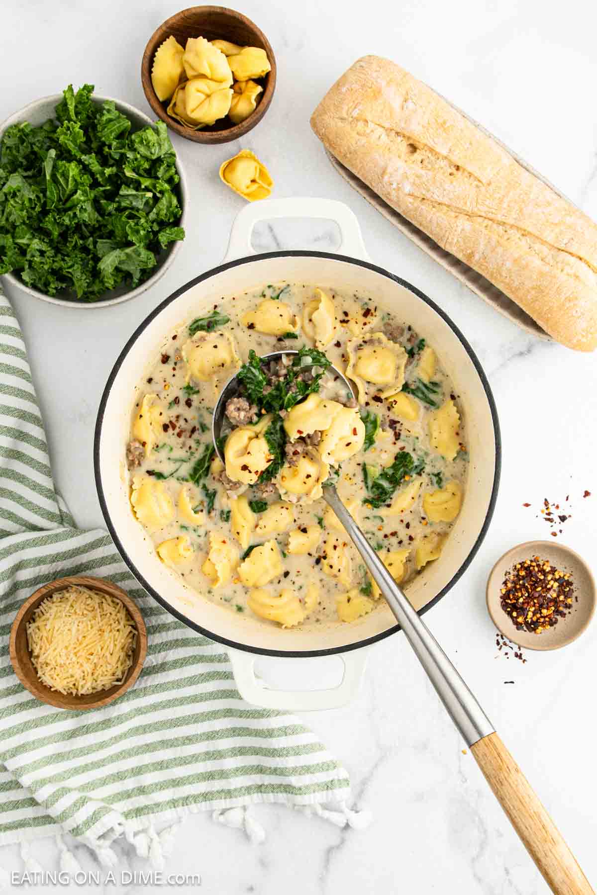 A pot of creamy sausage tortellini soup with kale sits next to a loaf of bread, a bowl of grated cheese, red pepper flakes, fresh kale, and a green-striped towel on a marble countertop.