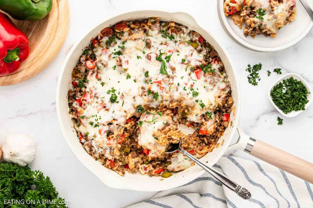 A skillet filled with cheesy Stuffed Pepper Casserole, topped with melted cheese and chopped parsley, sits on a white surface next to fresh peppers, garlic, and herbs. A serving spoon rests in the hearty casserole.