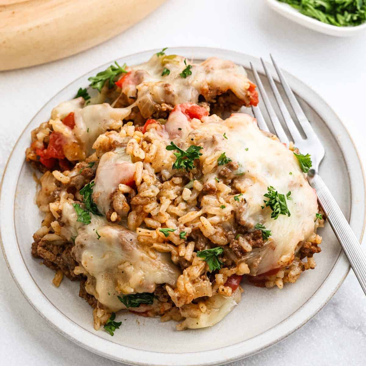 A plate of cheesy Stuffed Pepper Casserole, garnished with chopped parsley and served with a fork. This comforting casserole recipe features melted cheese, rice, ground beef, and bits of tomato for a hearty meal.