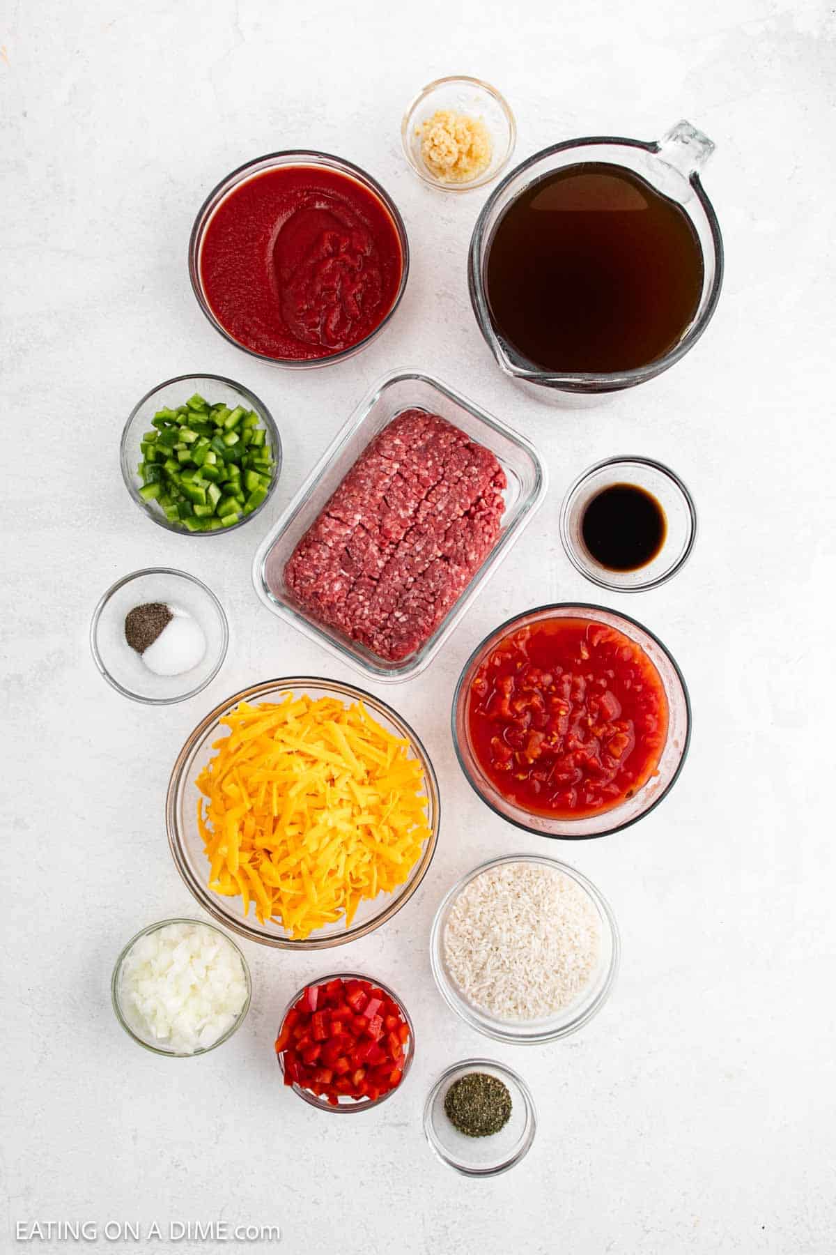 Top-down view of Stuffed Pepper Soup ingredients in bowls and containers, featuring ground beef, shredded cheddar cheese, diced tomatoes, broth, rice, bell peppers, onions, tomato sauce, garlic, and seasonings on a white surface.