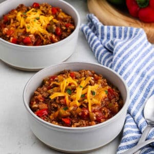 Two bowls of Stuffed Pepper Soup topped with shredded cheddar cheese sit on a light surface next to a blue-and-white striped towel, a spoon, and a wooden board with fresh bell peppers in the background.