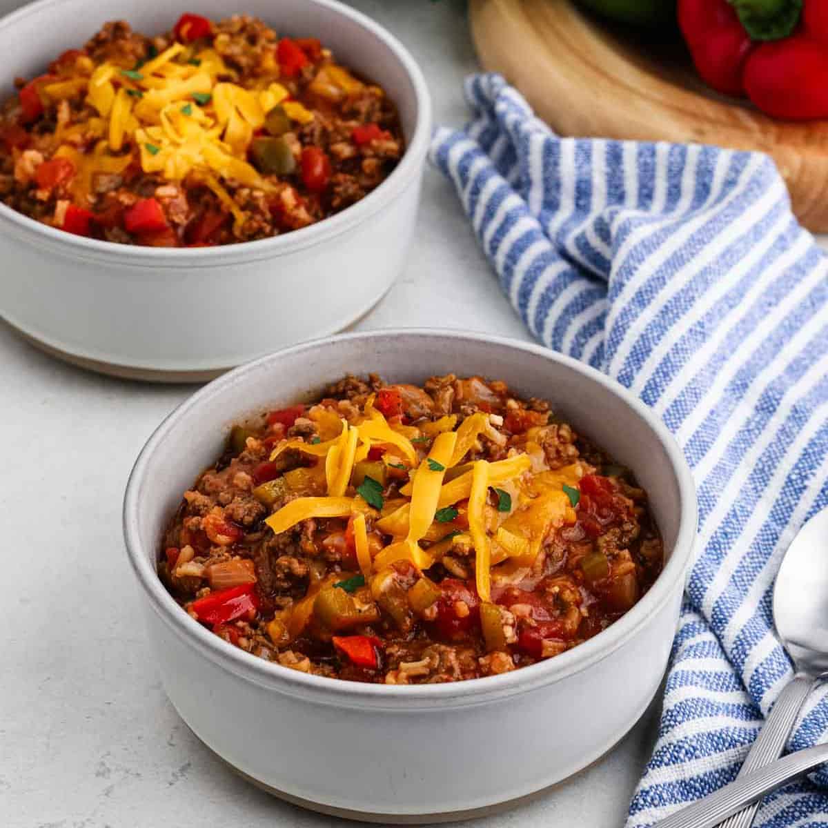 Two bowls of Stuffed Pepper Soup topped with shredded cheddar cheese sit on a light surface next to a blue-and-white striped towel, a spoon, and a wooden board with fresh bell peppers in the background.