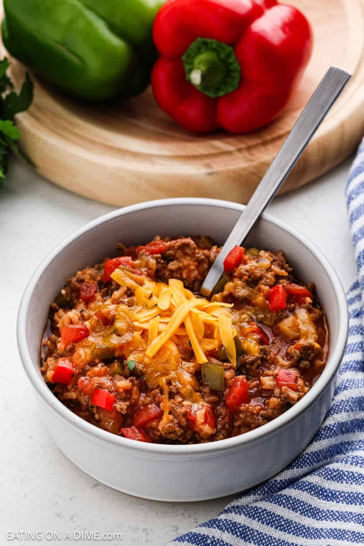 A bowl of Stuffed Pepper Soup topped with shredded cheese sits with a spoon inside. In the background, a whole red bell pepper and cutting board are visible, along with a blue and white striped cloth nearby.