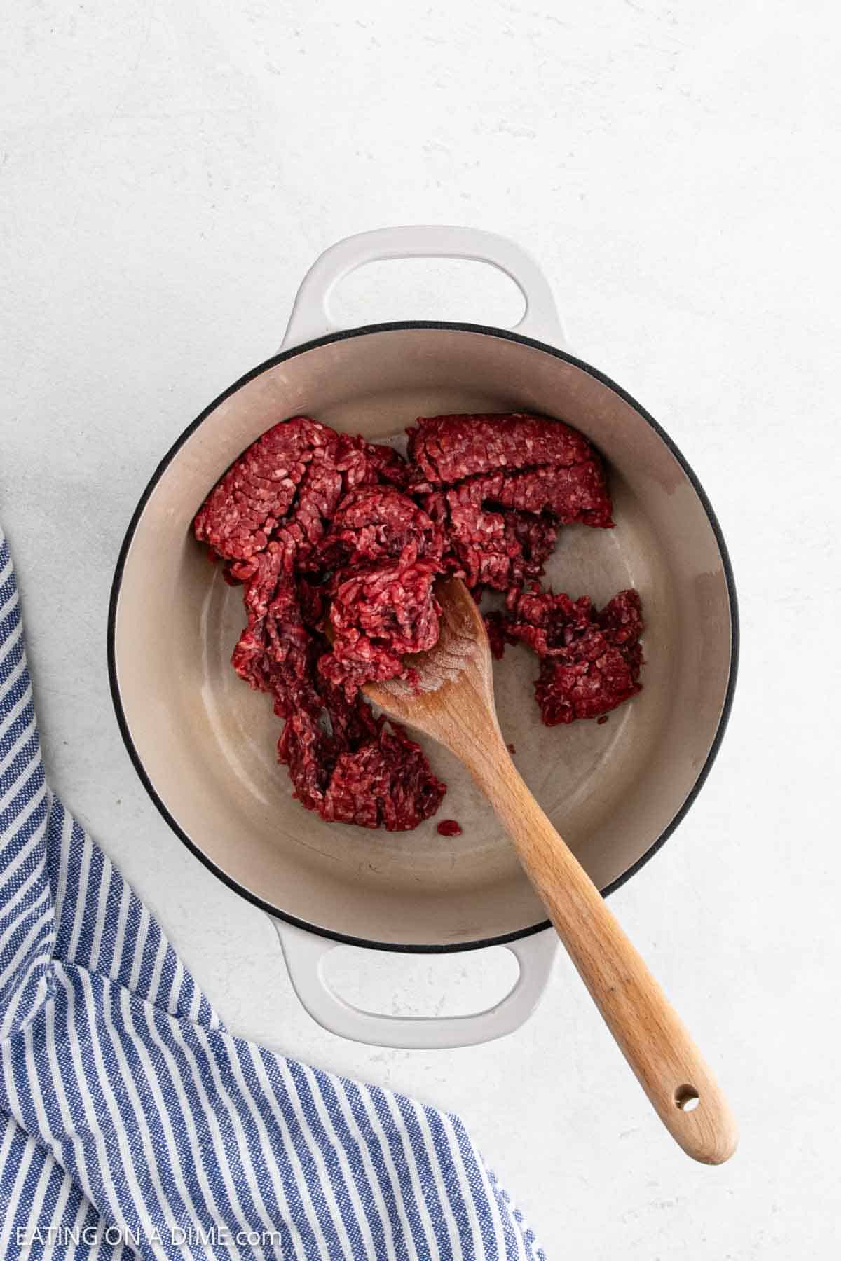 A white pot with raw ground beef being browned for Stuffed Pepper Soup, stirred with a wooden spoon. A blue and white striped kitchen towel is next to the pot on a light countertop.
