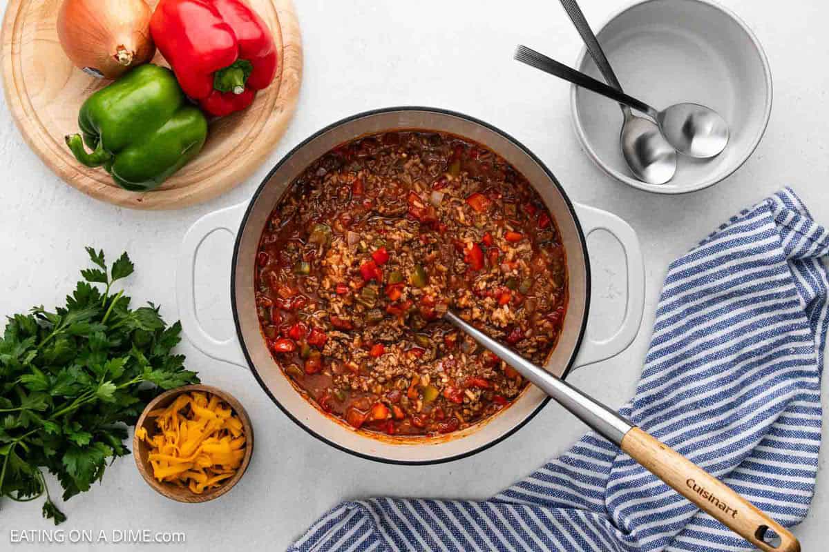 Overhead view of a pot filled with Stuffed Pepper Soup, surrounded by a bowl with spoons, a striped kitchen towel, chopped parsley, shredded cheese, and whole onions and bell peppers on a cutting board.