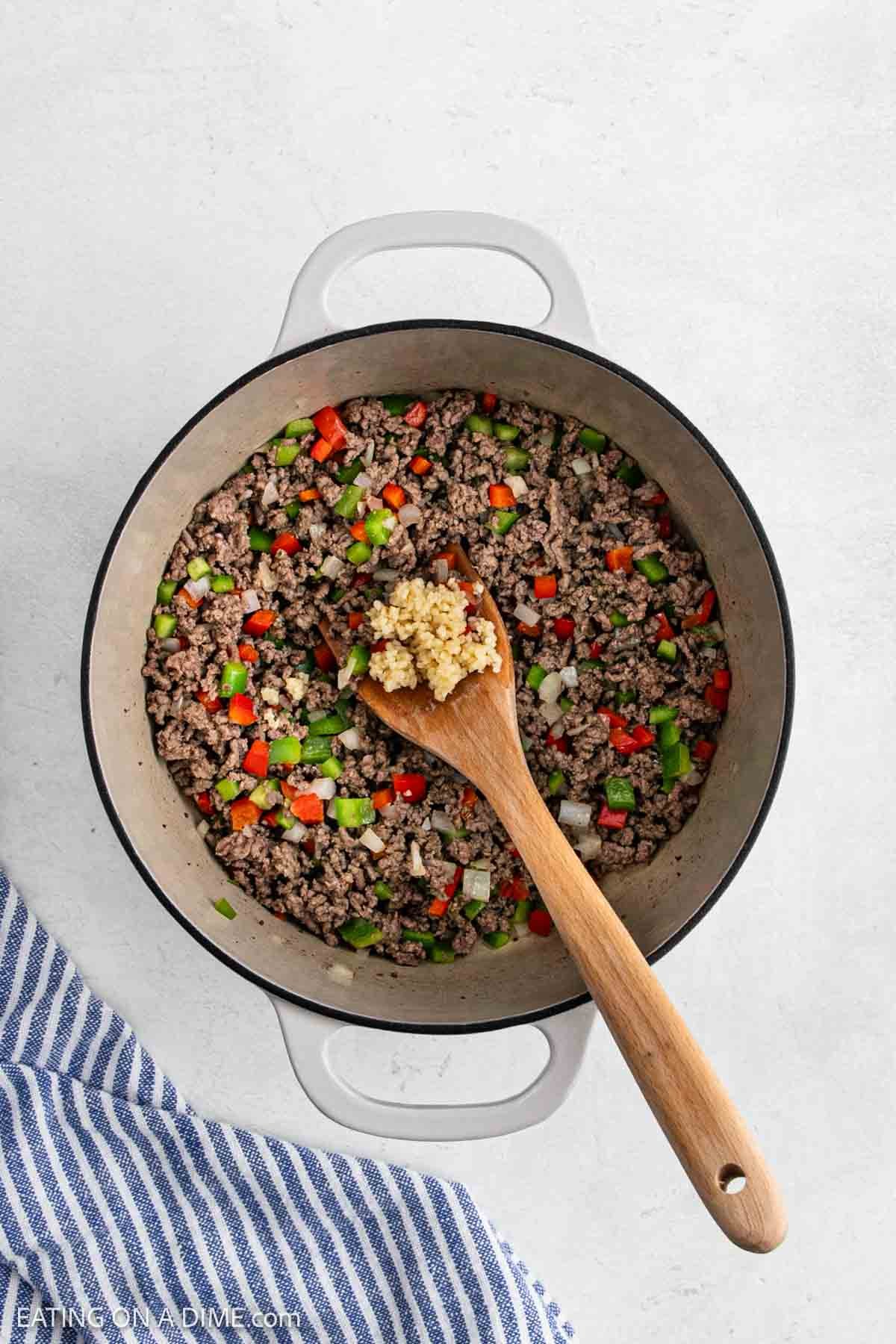 A pot filled with cooked ground beef, diced red and green bell peppers, and onions shows the beginnings of a hearty Stuffed Pepper Soup. A wooden spoon holding minced garlic rests over the mixture, with a blue and white striped towel beside the pot.