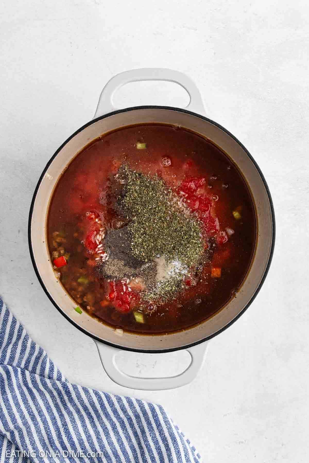 A white pot filled with Stuffed Pepper Soup ingredientsโchopped vegetables, diced tomatoes, and a generous pile of dried herbs and spices on topโsits on a light surface next to a blue and white striped towel.