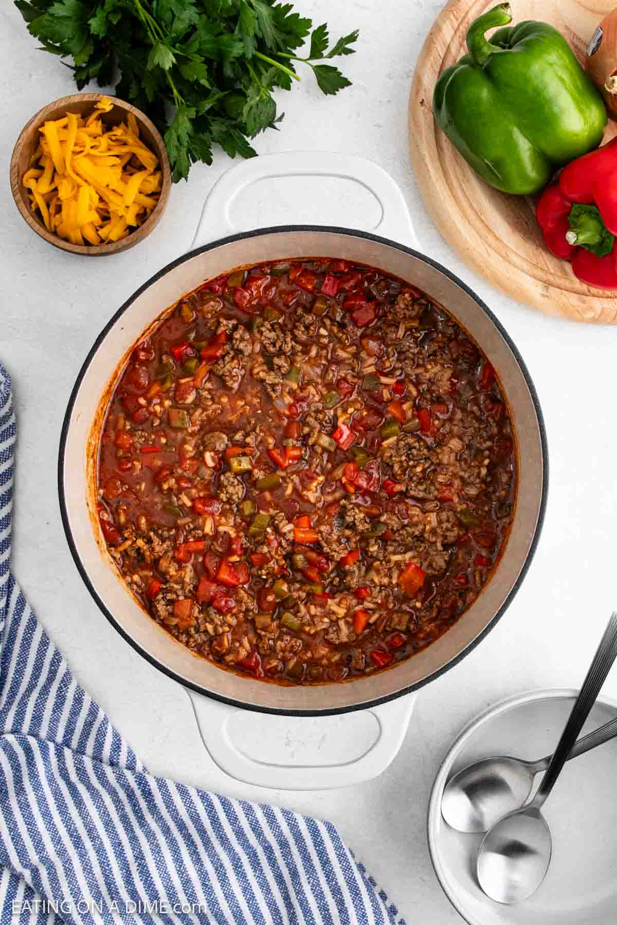 A pot of Stuffed Pepper Soup with ground meat, rice, and diced peppers sits on a white surface. Nearby are shredded cheese, fresh parsley, whole bell peppers, a striped towel, and two spoonsโperfect for this comforting soup recipe.