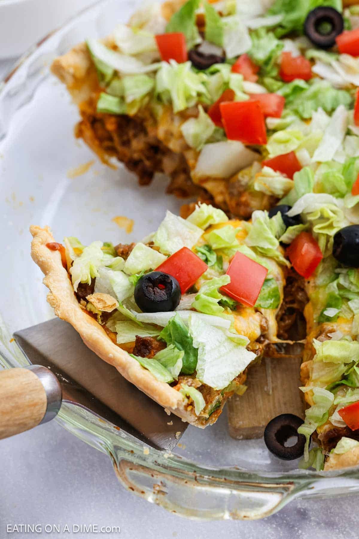 A slice of taco pie topped with shredded lettuce, diced tomatoes, chopped onions, and black olives is being lifted from a glass pie dish. The filling and toppings are colorful and fresh.
