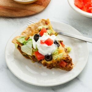 A slice of taco pie topped with shredded lettuce, diced tomatoes, black olives, and a dollop of sour cream sits on a white plate with a fork.