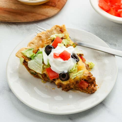 A slice of taco pie topped with shredded lettuce, diced tomatoes, black olives, and a dollop of sour cream sits on a white plate with a fork.