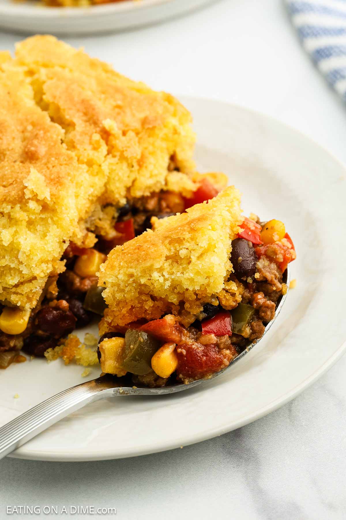 A close-up of a plate with cornbread casserole, showing a spoonful lifted. The casserole has a golden cornbread topping with a filling of ground beef, black beans, corn, tomatoes, and bell peppers.