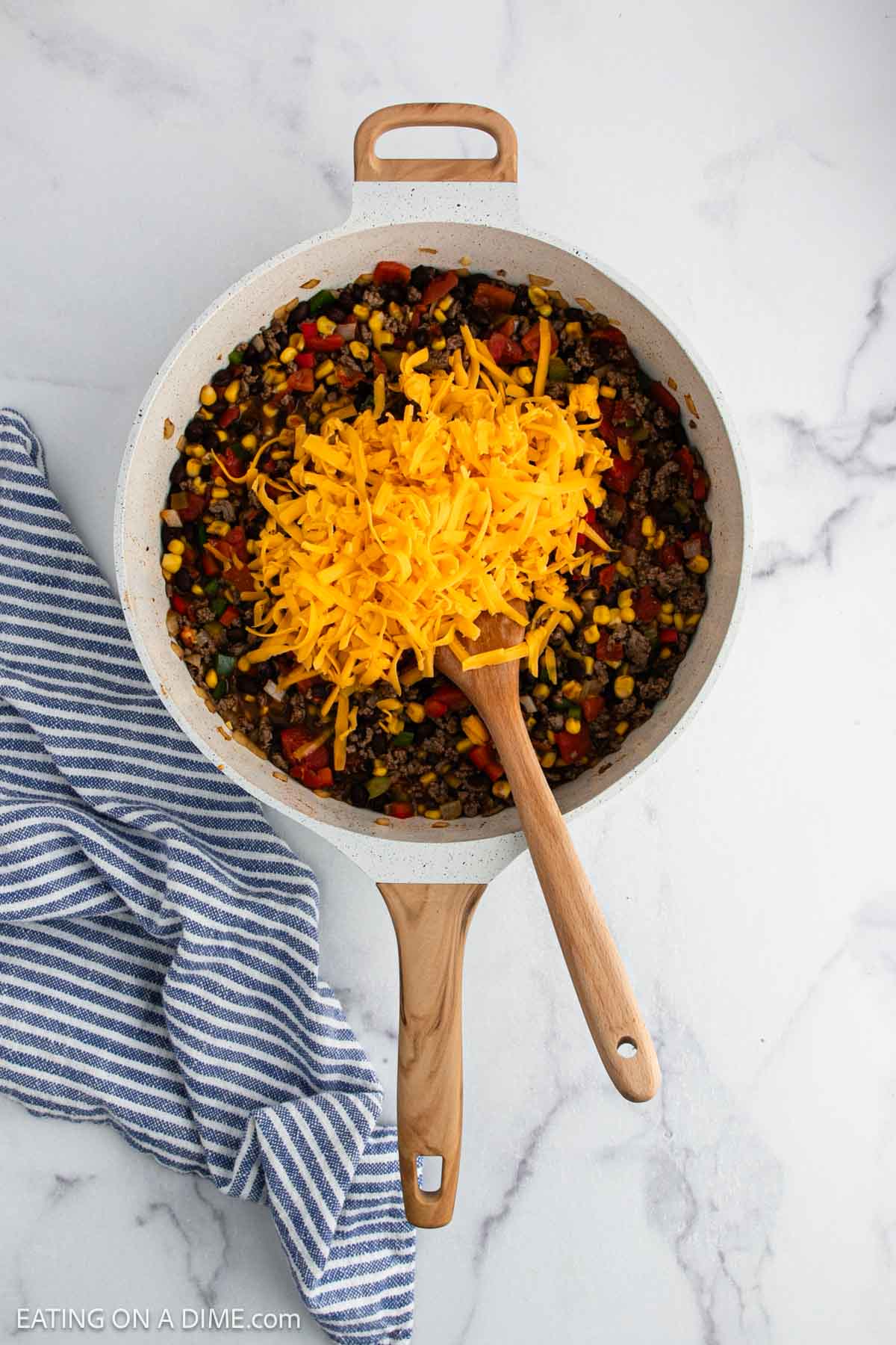 A skillet filled with a mixture of black beans, corn, and bell peppers, topped with a pile of shredded cheddar cheese. A wooden spoon rests inside the pan, and a striped towel lies beside it on a marble surface.