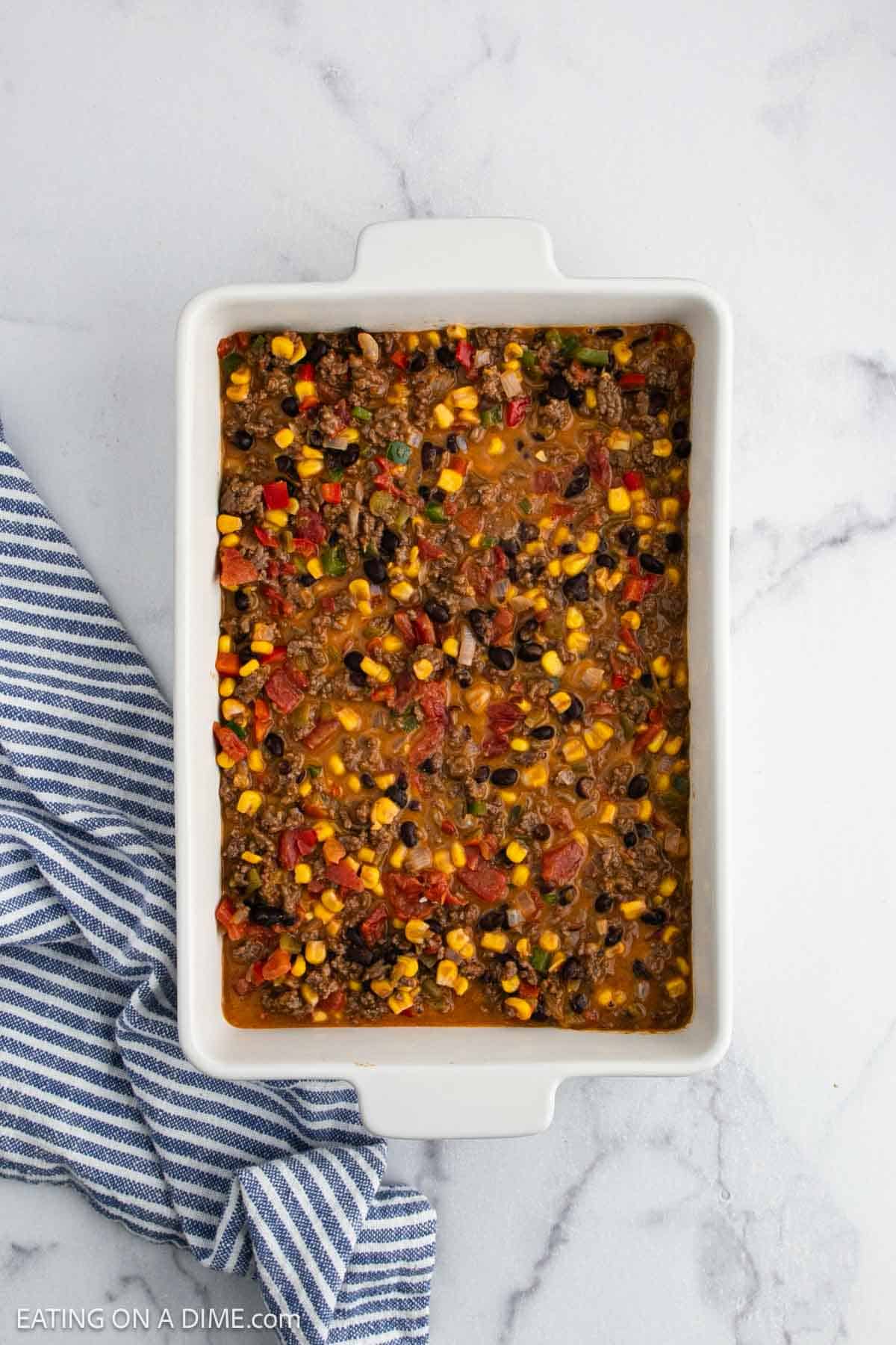 A rectangular white baking dish filled with a colorful mixture of ground meat, black beans, corn, tomatoes, and peppers sits on a marble surface beside a blue and white striped towel.