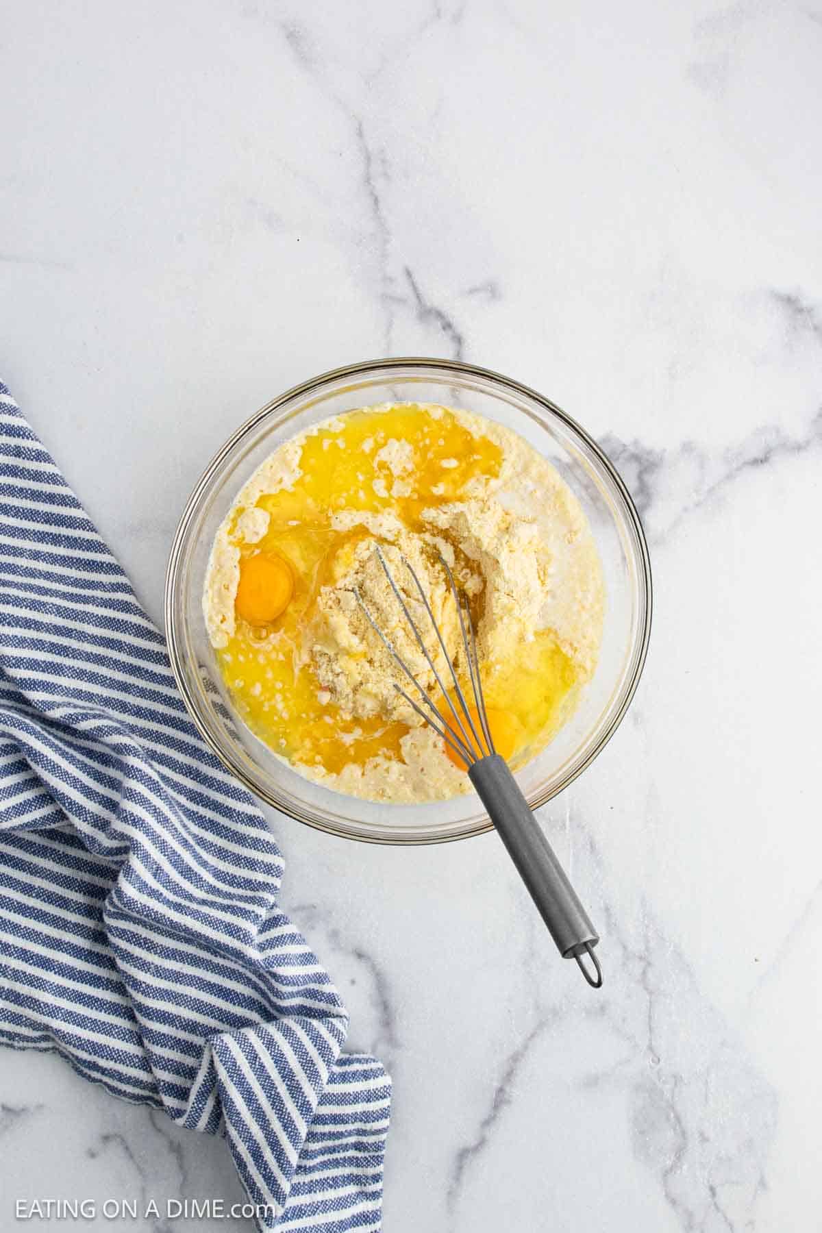 A glass bowl filled with flour, eggs, and liquid ingredients sits on a marble surface with a whisk inside. A blue and white striped towel is nearby.