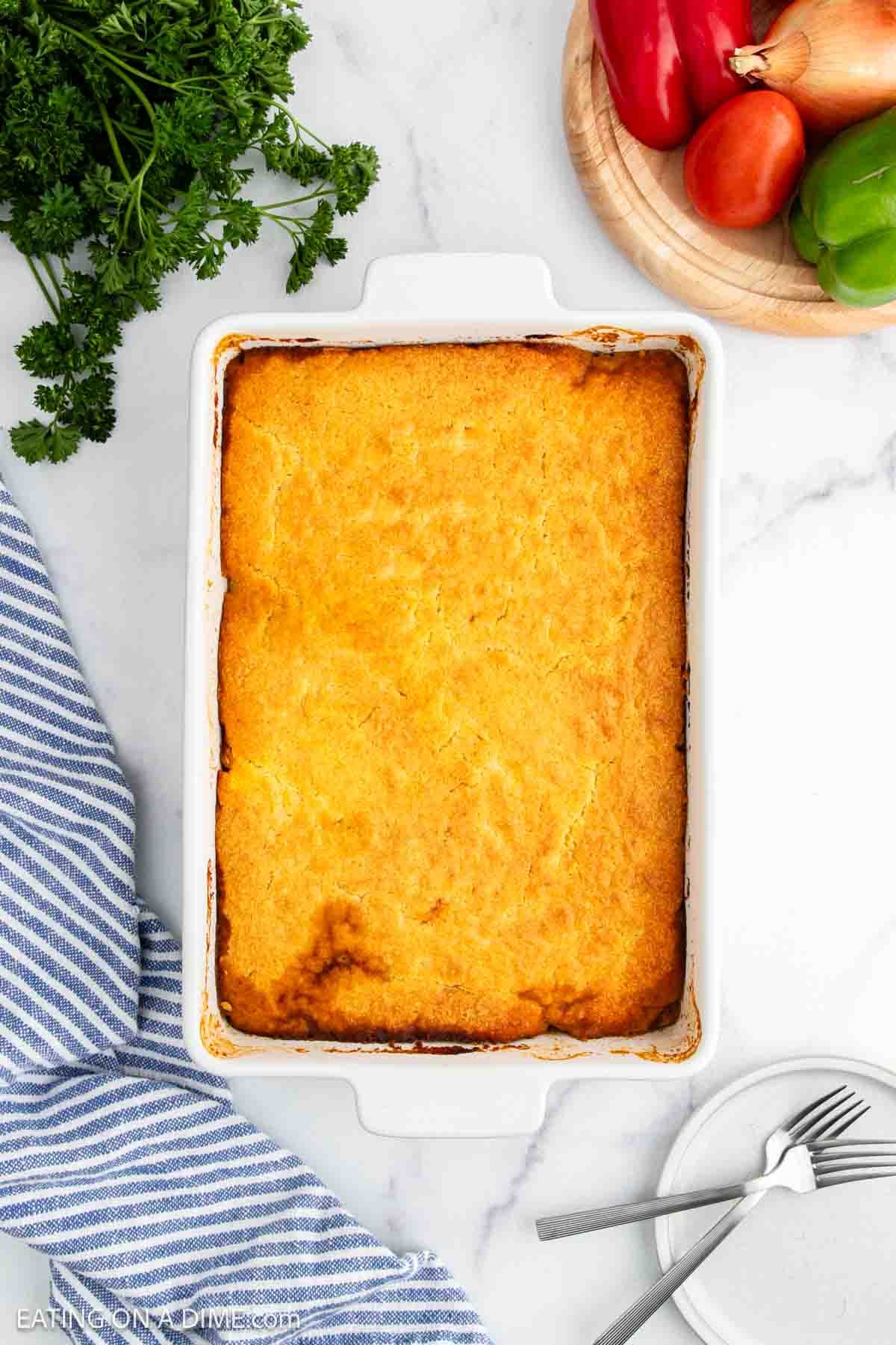 A baked casserole with a golden, slightly browned top in a white rectangular dish, surrounded by parsley, fresh vegetables, a striped towel, and a plate with forks on a marble surface.