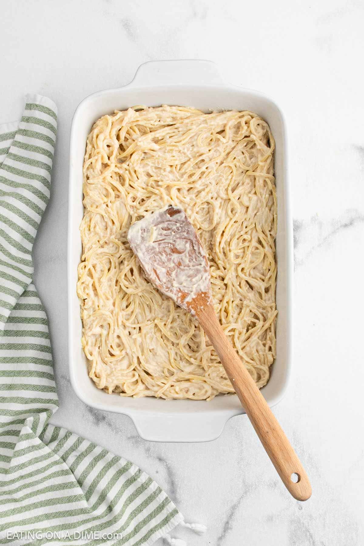 A white baking dish filled with creamy Alfredo Spaghetti, topped with a wooden spatula coated in sauce. A green and white striped towel rests nearby on the white marble surface—perfect for trying the latest TikTok recipes at home.
