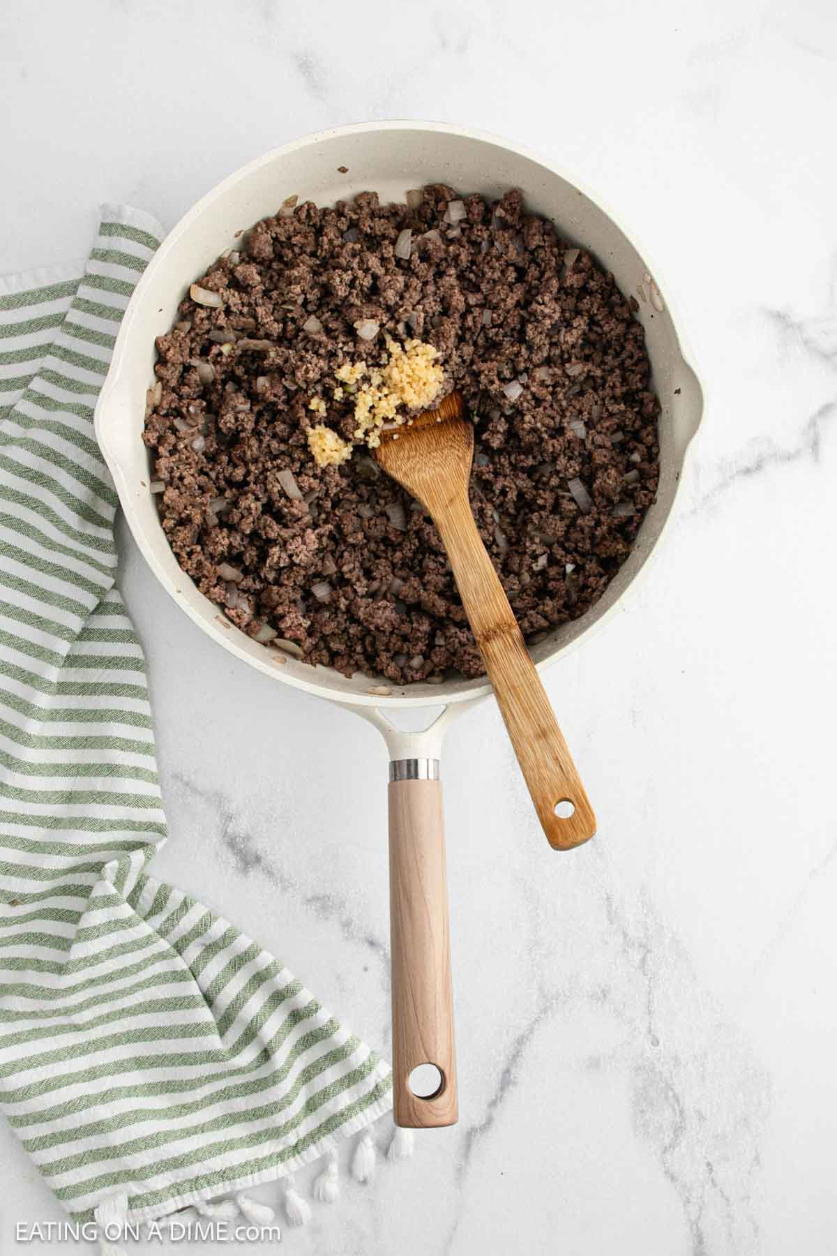 A skillet with cooked ground beef and chopped onions, a wooden spoon resting inside, and a small pile of minced garlic on top—perfect for starting your own TikTok Alfredo or TikTok pasta. A green and white towel sits beside the skillet on a marble surface.